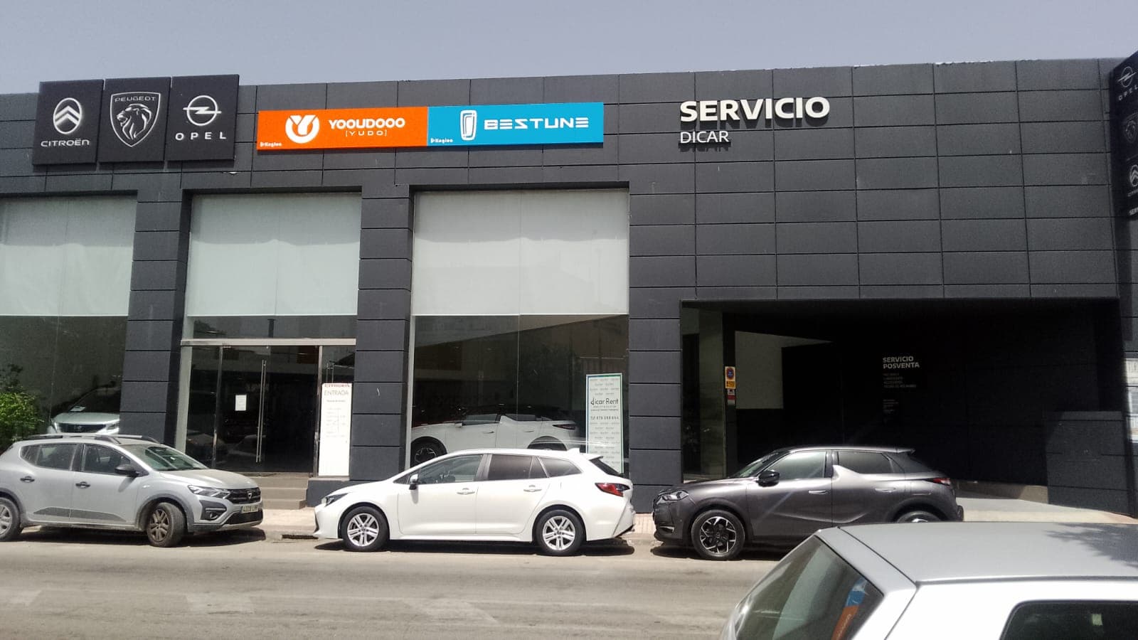 Fachada de concesionario multimarca con logotipos de coches y varios vehículos estacionados enfrente.