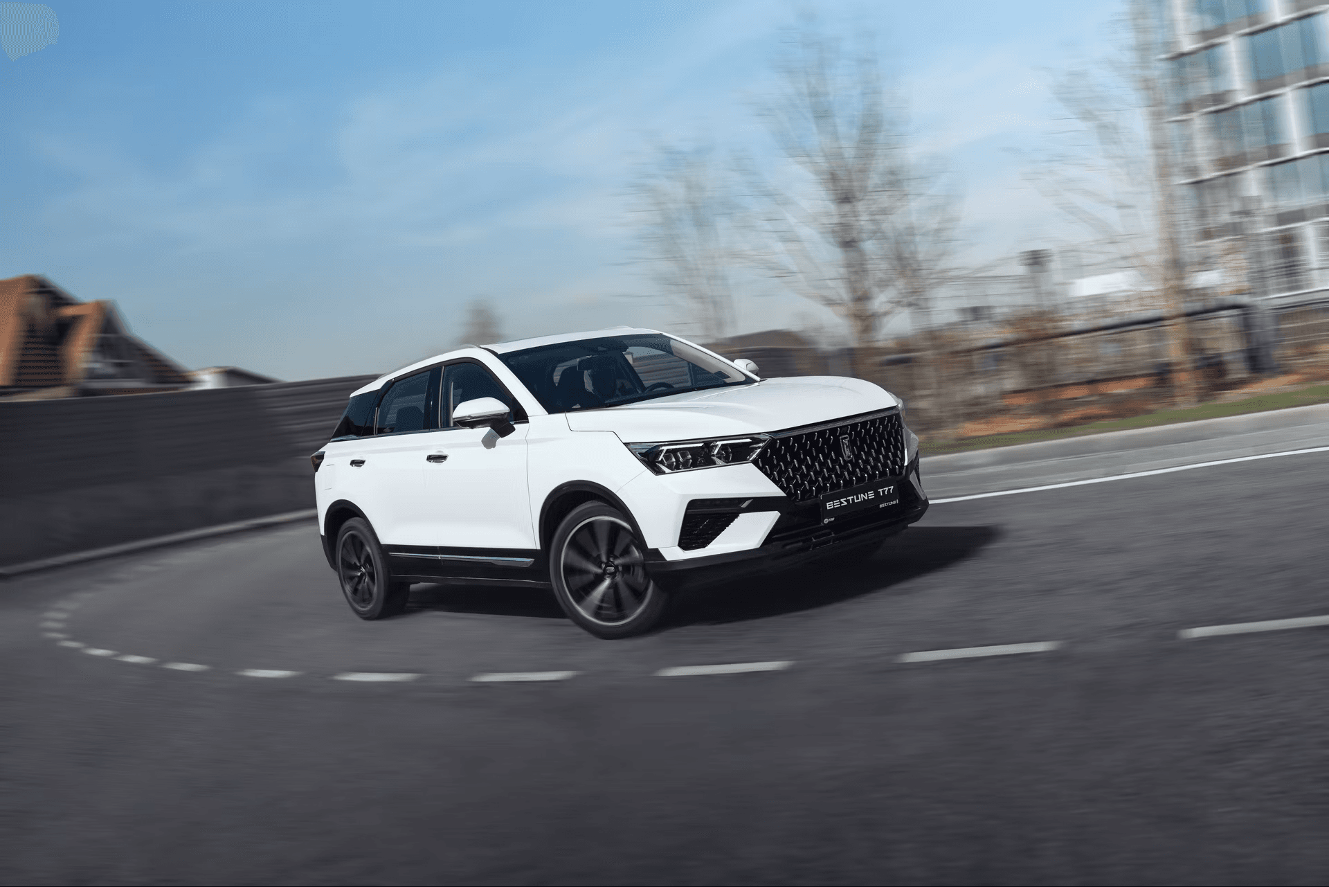 SUV blanco Bestune T77 circulando velozmente por una carretera curva con el fondo urbano desenfocado.