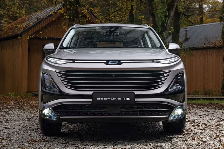 Vista frontal de un SUV Bestune T90 plateado estacionado en grava con cabañas de madera.